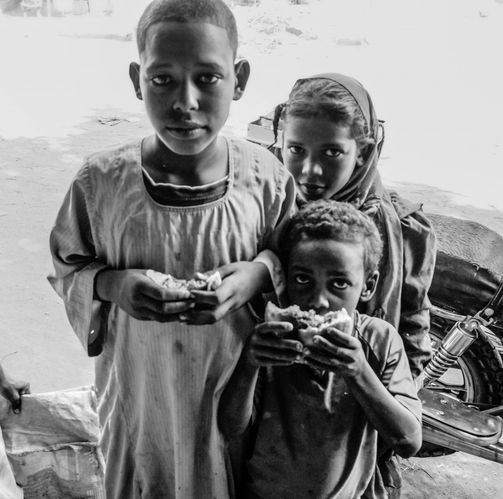 Bambini profughi nel villaggio di Kerma, in gruppo vicino ai banchetti di cibo per strada aspettando che qualcuno gli lasci qualcosa, qui mostrano un panino che gli è stato offerto. Foto di Angelo Calianno.